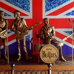 Pomnik The Beatles na tle flagi Wielkiej Brytanii. Czterech muzyków w pozłacanych strojach, z gitarami i perkusją, upamiętnia legendę muzyki.