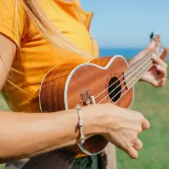 Dziewczyna w pomarańczowej bluzce gra na ukulele na tle morza i zielonej łąki.