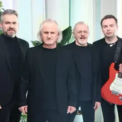 Czterech mężczyzn w czarnych strojach. Jeden z nich trzyma czerwoną gitarę, gotową do wykonania utworów.