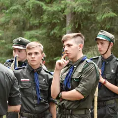 Harcerze w mundurach czekają na rozpoczęcie modlitwy obozowej. W lesie panuje skupienie.