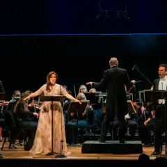 Trzech śpiewaków operowych, w tym kobieta w złotej sukni, wykonuje wzruszającą arię z towarzyszeniem orkiestry symfonicznej. Dyrygent kieruje wykonaniem.