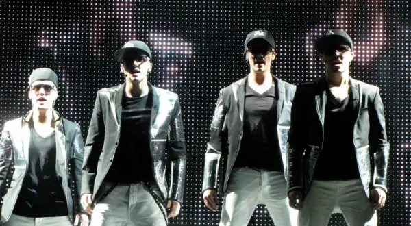Członkowie Big Time Rush w błyszczących marynarkach i czapkach z daszkiem, na tle sceny z migoczącymi światłami.