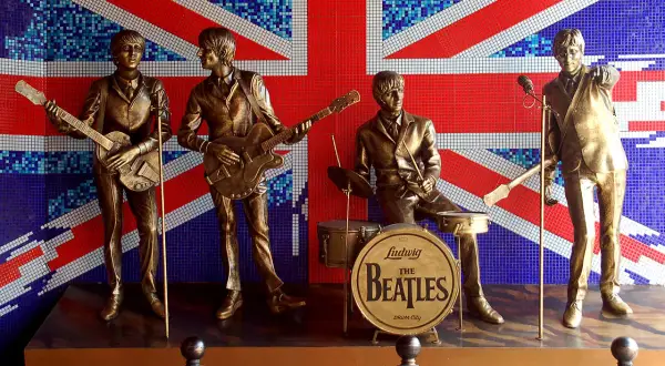 Pomnik The Beatles na tle flagi Wielkiej Brytanii. Czterech muzyków w pozłacanych strojach, z gitarami i perkusją, upamiętnia legendę muzyki.