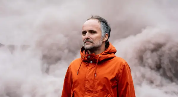 Artur Rojek w pomarańczowej kurtce, na tle dymu. Jego twórczość często porusza tematykę przemijania i refleksji.