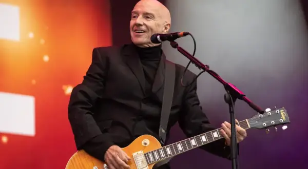 Łysy mężczyzna w czarnym garniturze gra na gitarze elektrycznej, uśmiechając się. To jest jego chwila, bez żalu.