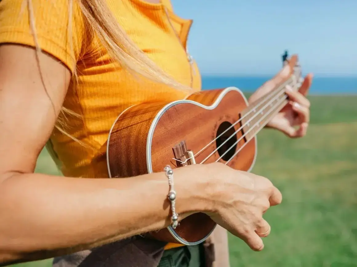 Dziewczyna w pomarańczowej bluzce gra na ukulele na tle morza i zielonej łąki.