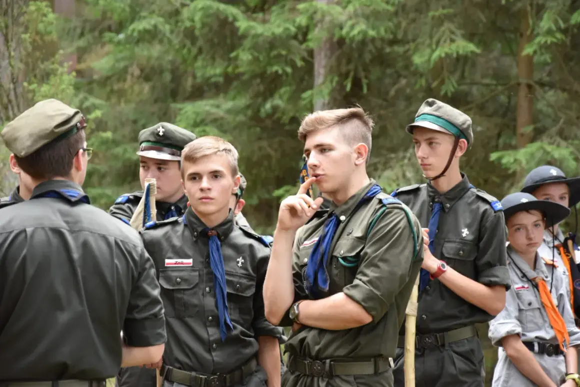 Harcerze w mundurach czekają na rozpoczęcie modlitwy obozowej. W lesie panuje skupienie.