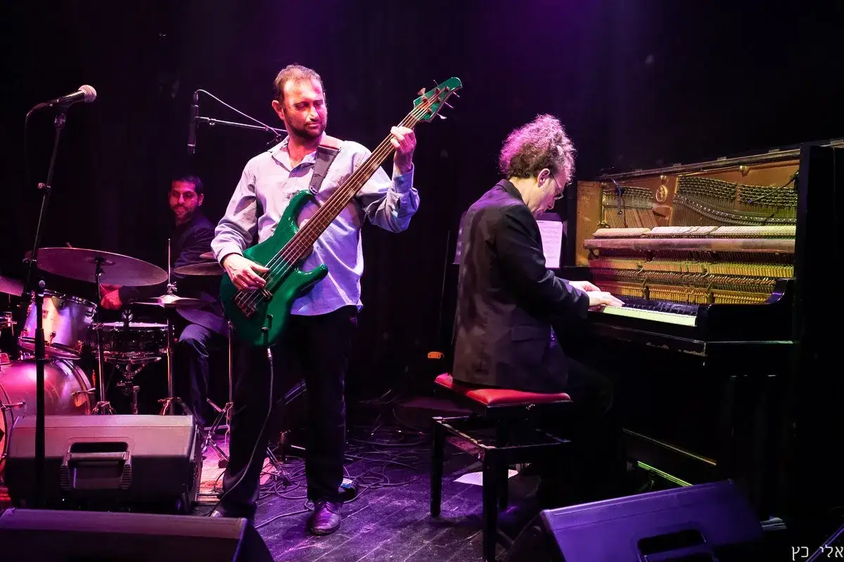 Muzycy grają jazzową balladę. Basista z zieloną gitarą, pianista i perkusista tworzą nastrojową atmosferę.