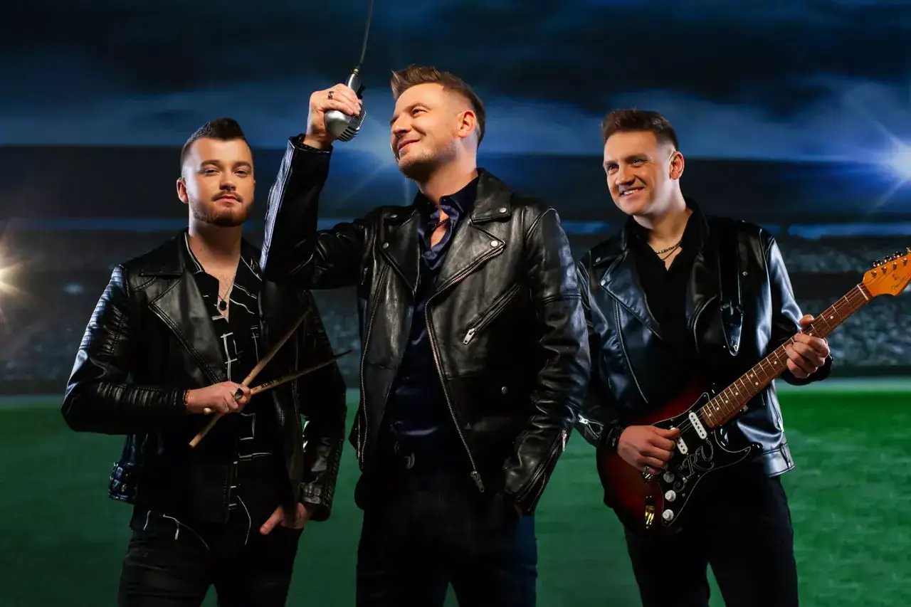 Trzech mężczyzn w skórzanych kurtkach, jeden z mikrofonem, drugi z gitarą, trzeci z pałeczkami. Jakby grali koncert w maju w tramwaju.