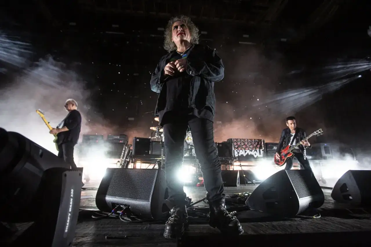 Robert Smith z The Cure na scenie, otoczony dymem i światłami. Muzyka zespołu The Cure brzmi potężnie.